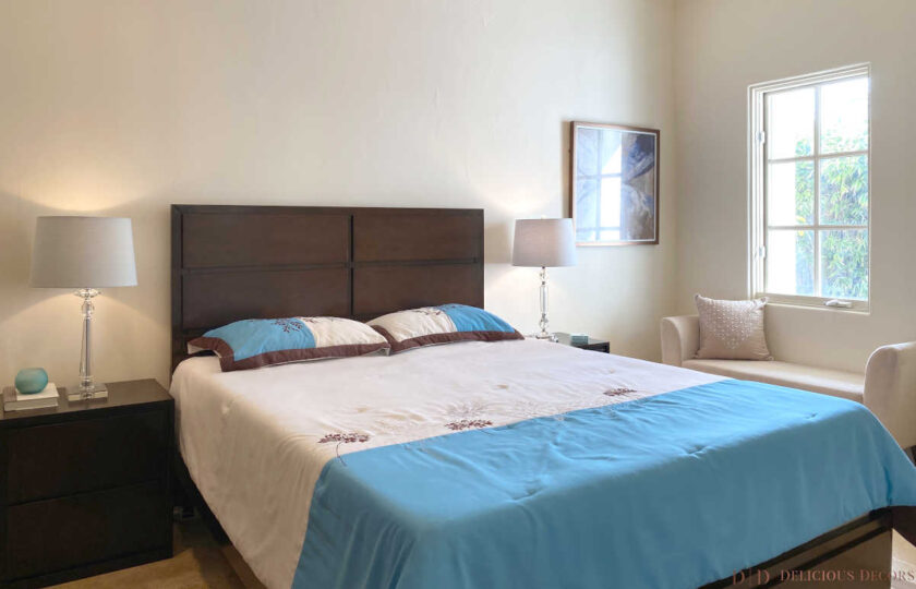 Guest bedroom facing queen bed with brown traditional wood headboard, blue and white bedding, and fabric benche to the right