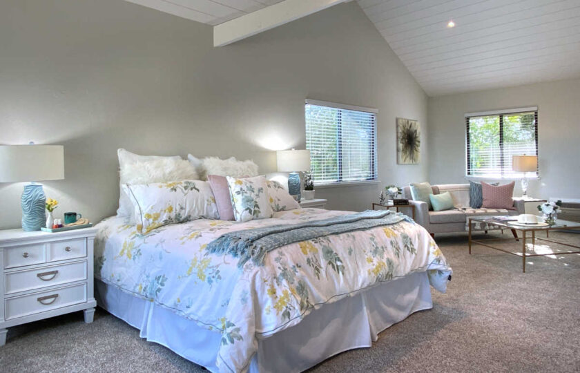 Master bedroom with flower bedding, white wood nightstands, and love seat setup in the background