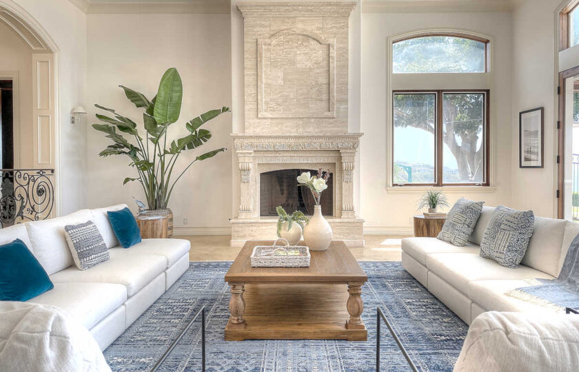 White matching sofas and two contemporary armchairs surround a grand fireplace in home staging a modern traditional Malibu compound. A large palm is in the corner and a traditional oak coffee table is in the center.