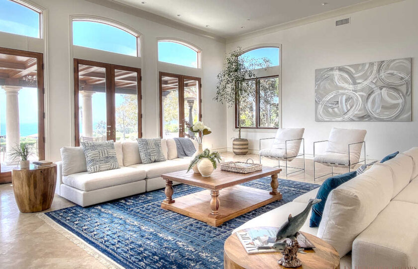This is an angled view of white matching sofas and two modern club chairs surrounding a grand fireplace in a Malibu home. A large palm is in the corner and a traditional oak coffee table is in the center.