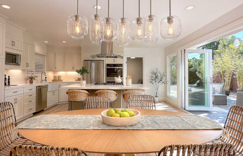 The edison bulb chandelier sits over a starburst oak dining table, looking to the open-area remodeled kitchen.