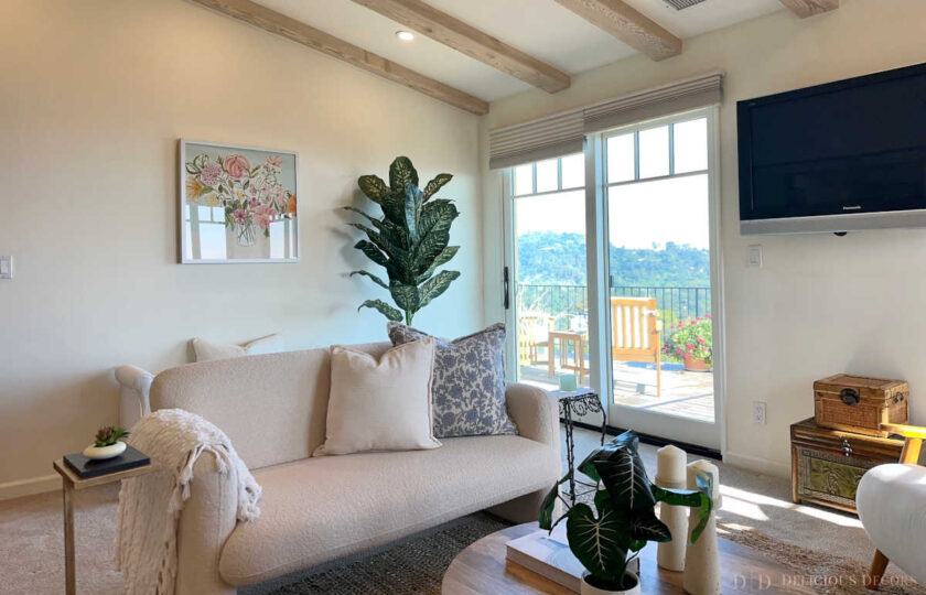 Cream bouclé sofa with a blue botanical pillow and pastel floral art in a modern Mediterranean den with exposed wood beams.