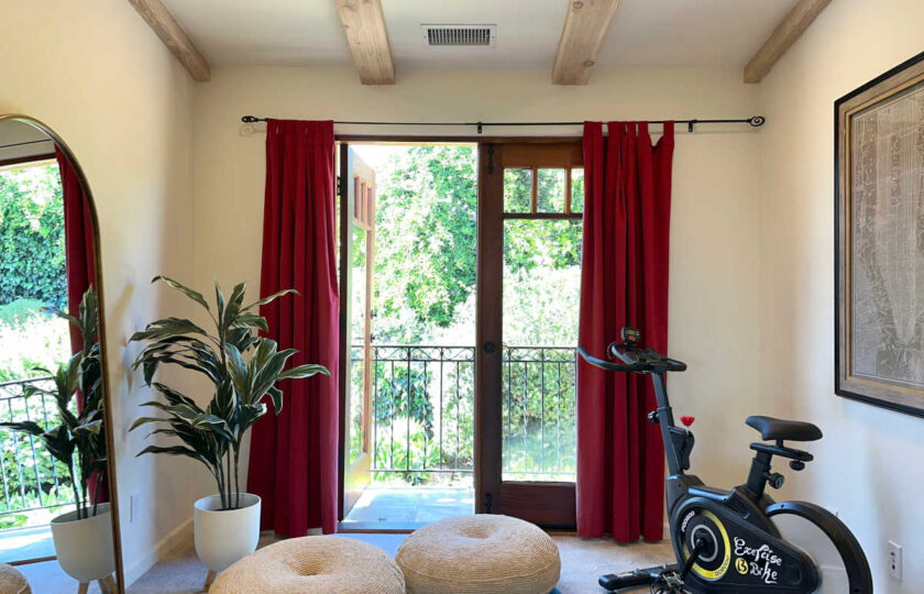 Wellness studio with stationary bike, woven poufs, mirror, and red curtains over French doors leading to a lush balcony.