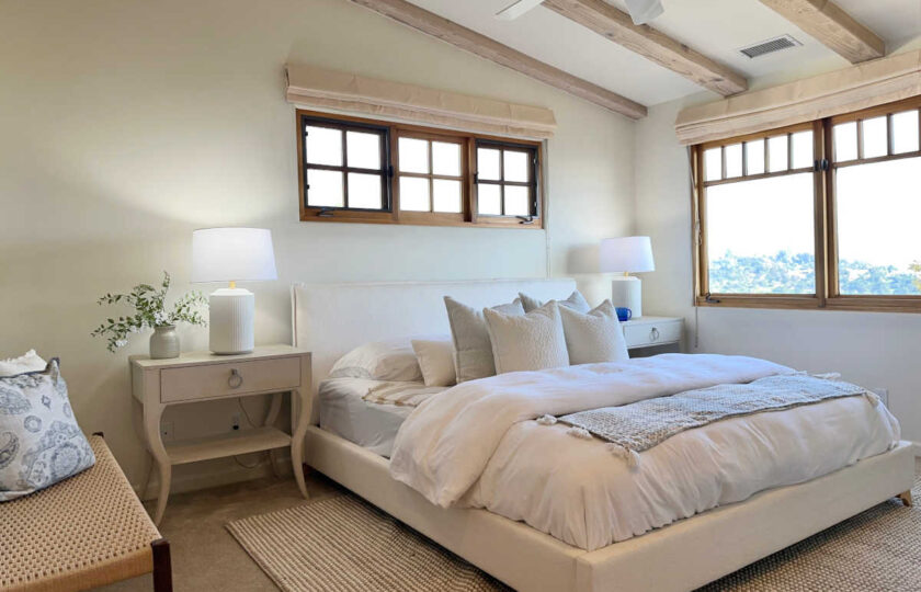 Serene primary suite showing exposed wood beams, a white upholstered bed, textured rug, and custom windows with hillside view.