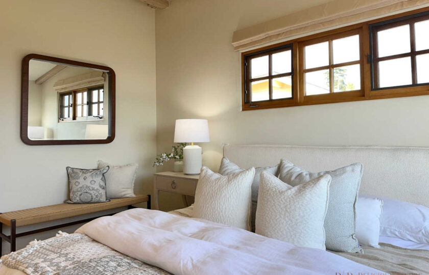 Mediterranean primary bedroom featuring a white upholstered bed with textured linens, woven bench, and wood-framed windows.
