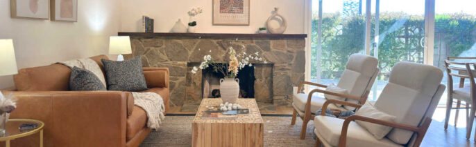 West Pasadena home staging featuring a tailored tan leather sofa, raw block-wood coffee table, and wood-framed accent chairs in front of a classic stone fireplace.