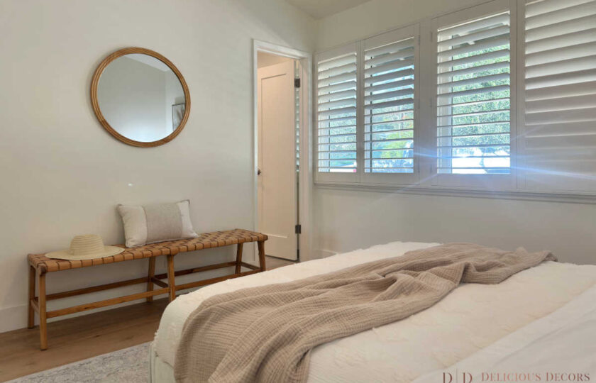 Calm bedroom retreat styled with a woven leather bench, layered neutral muslin bed throws, and bright white plantation shutters.