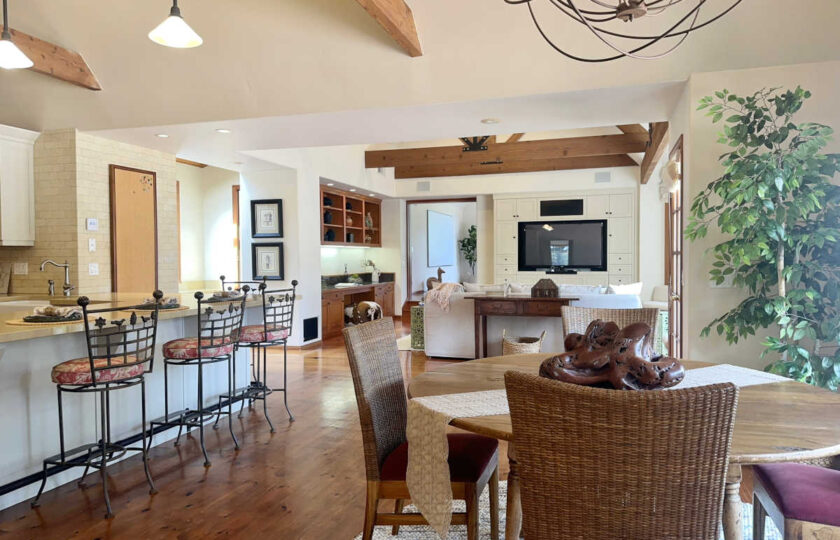 Open-concept dining and kitchen area staged with a round wood dining table, woven dining chairs, and rustic iron barstools under exposed timber beams.