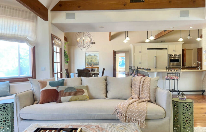 Open-concept family room staging featuring a light linen sofa and rust cotton pillows looking past a woven kilim ottoman into a bright, modern kitchen.