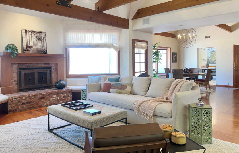 Highland cabin style family room showcasing a woven kilim ottoman, a light linen sofa with soft rust cotton throws, and a red brick fireplace under vaulted timber trusses.