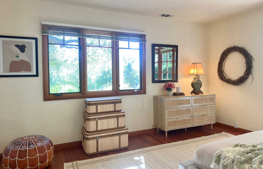 Santa Ynez luxury estate staging in the bright, botanical "hers" room featuring a woven rattan dresser, complemented by stacked decorative trunks and a stitched leather pouf.