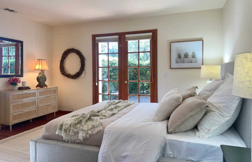 Santa Ynez luxury estate staging in a "hers" guest retreat , featuring a grey bed, a woven rattan dresser, and glass French doors leading to the lush grounds.