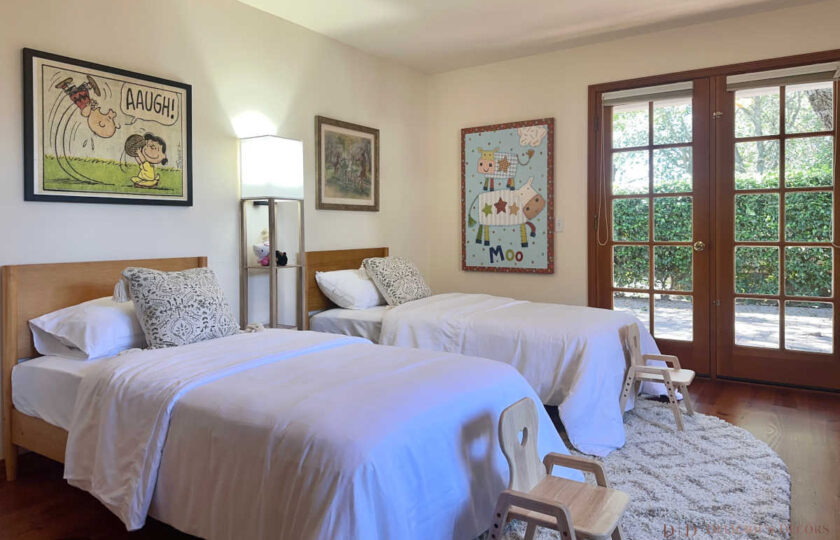 Playful children's room in a Santa Ynez luxury estate staging, anchored by blonde wood twin beds, soft white bedding, and whimsical art near glass doors.