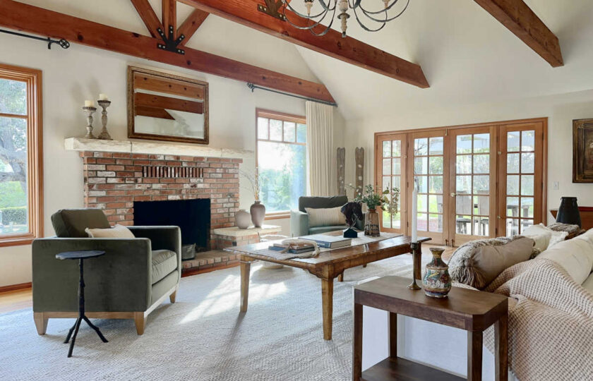 Formal living room staged with a rustic luxury aesthetic, showcasing a red brick fireplace, an oversized wood coffee table, and olive green armchairs looking toward glass French doors.