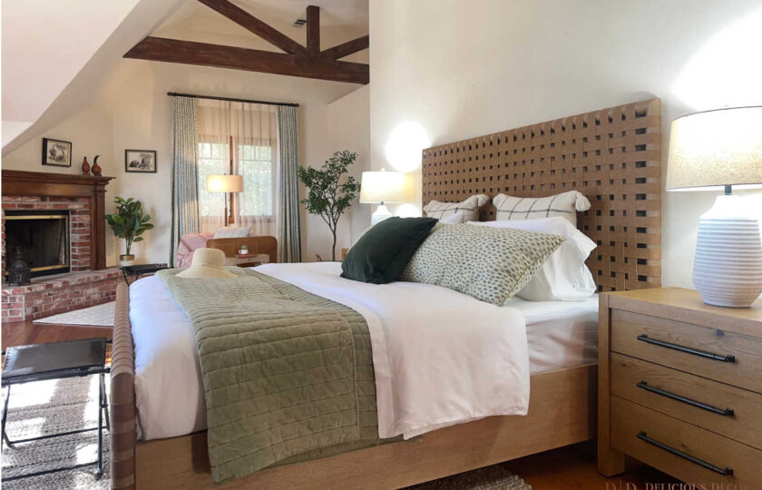 Close-up of primary suite staging showing a woven cognac leather headboard layered with textured green bedding and light linen pillows next to a classic red brick fireplace.