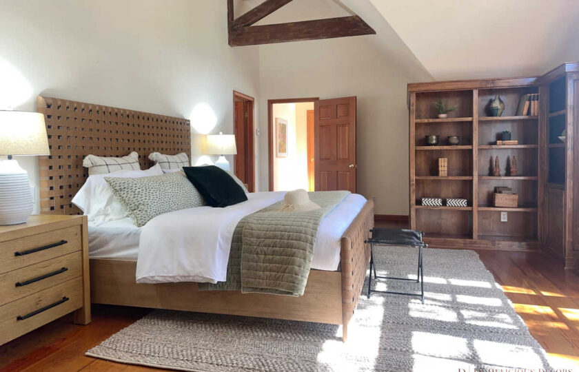 Santa Ynez luxury estate staging in a vaulted primary bedroom, anchored by a woven cognac leather headboard, a heavy wooden nightstand, and a built-in bookshelf.