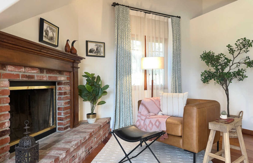 Primary suite sitting area warmed up by a caramel leather armchair tucked by a private red brick fireplace with a wood mantel.