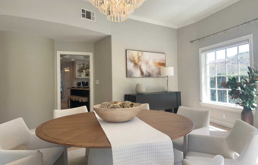 Staged formal dining room featuring large round wood dining table, white upholstered chairs, crystal chandelier, and dark sideboard.