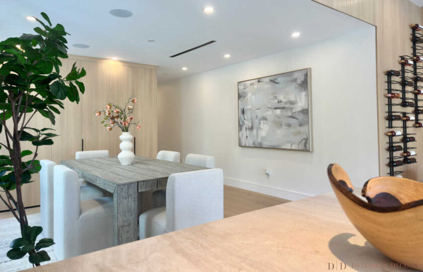 Staged dining room showcasing a rustic gray wood table with cream upholstered chairs looking toward an open kitchen island with travertine countertops.