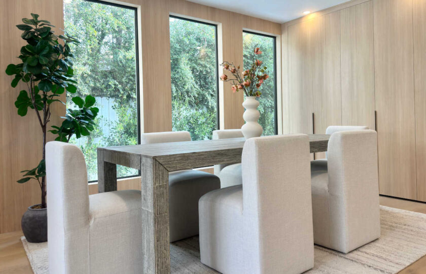 Dining area staged with natural light, large windows, light oak paneled walls, and a tall potted green floor plant.