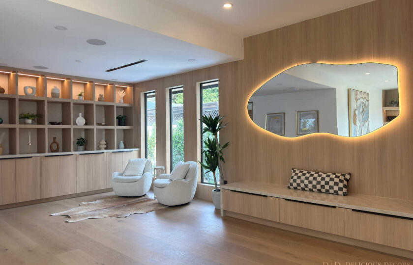 Staged entryway lounge showcasing an organic backlit wall mirror, custom light oak cabinetry, a cowhide rug, and white bouclé swivel armchairs.