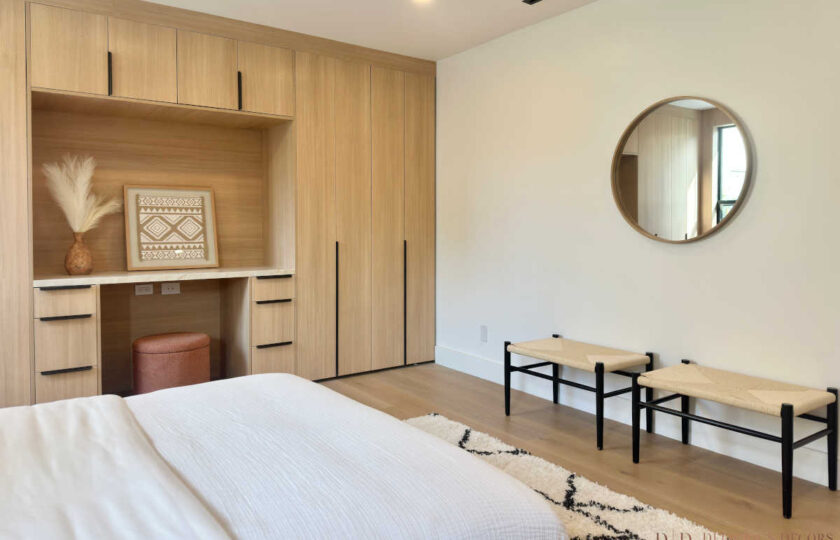 Bedroom suite featuring light oak built-in cabinetry, a vanity desk nook, woven accent benches, and a round wall mirror.