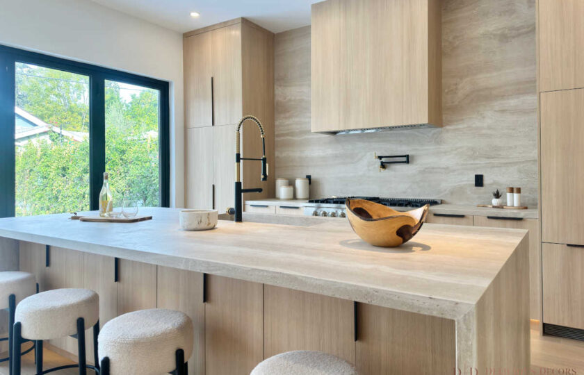 Modern organic kitchen staging featuring a travertine waterfall island, white bouclé counter stools, black hardware, and a custom wood range hood.