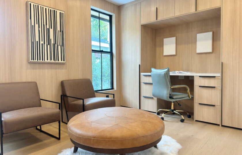 Dedicated home office space staged with light oak built-in desk cabinetry, a leather executive swivel chair, and a round tan leather ottoman.