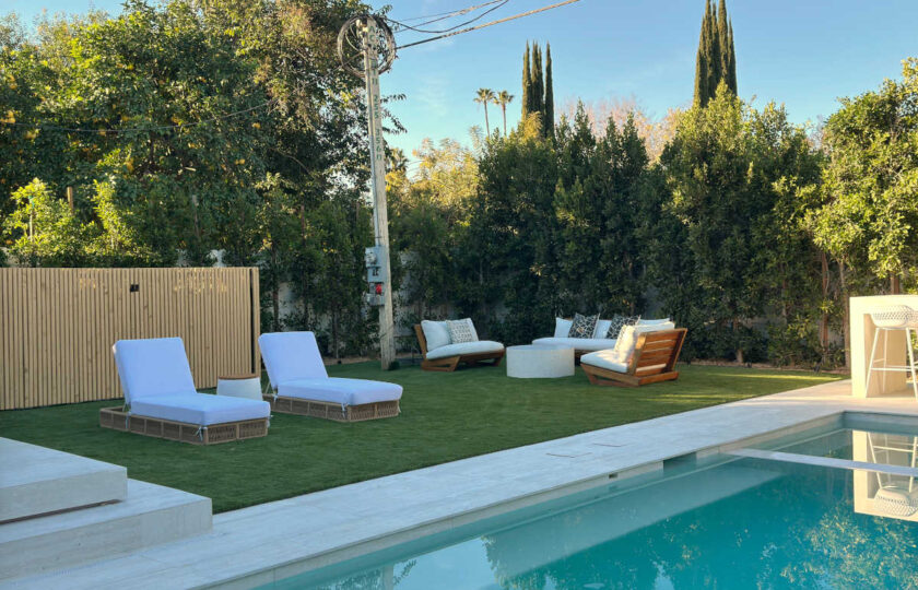 Backyard pool area staged with modern wood lounge chairs, outdoor sofas, a circular stone coffee table, and green turf.