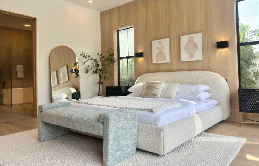 Primary bedroom suite view showcasing a arched floor mirror, light oak paneled wall, potted tree, and en-suite bathroom entry.