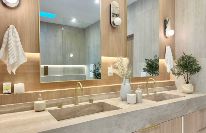 Luxury primary bathroom staging featuring double travertine vessel sinks, brass fixtures, marble sconces, and custom oak cabinetry.