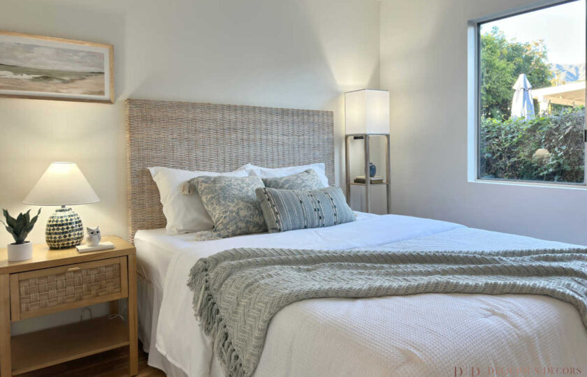 Guest bedroom corner view showing a patterned mosaic table lamp, wicker nightstand, and woven rattan headboard with soft blue-grey textiles.
