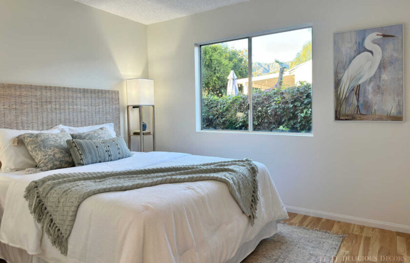 Coastal guest bedroom staging featuring a woven rattan headboard bed, blue and white patterned bedding, tall floor lamp, and heron bird artwork.