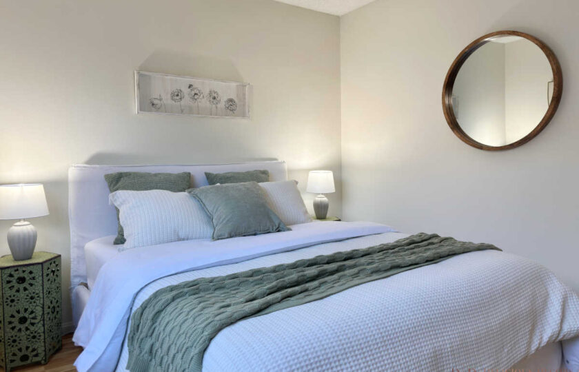 Staged bedroom angle featuring a round wood-framed wall mirror, white bed, green accent pillows, and decorative bedside table.