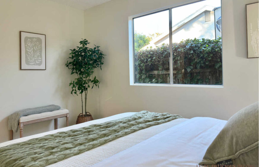 Cozy bedroom corner styled with a wooden bench, sage throw blanket, potted ficus tree, and window view of the outdoor hedge.