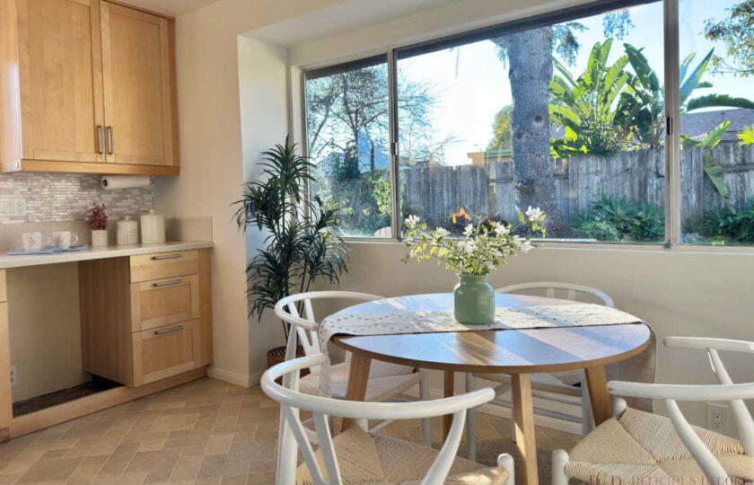Bright kitchen dining nook styled with a round oak table, white wishbone chairs, sage vase with white florals, and large window overlooking garden trees.