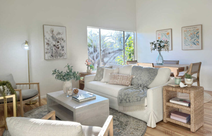 California Casual open-concept living and dining space styled with a white slipcovered sofa, light oak dining set, woven seagrass end table, and botanical wall art.