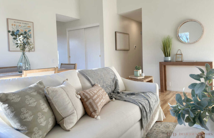 Sunlit living space styled with a neutral white sofa, sage and tan textured pillows, blue throw blanket, and entry console in the background.