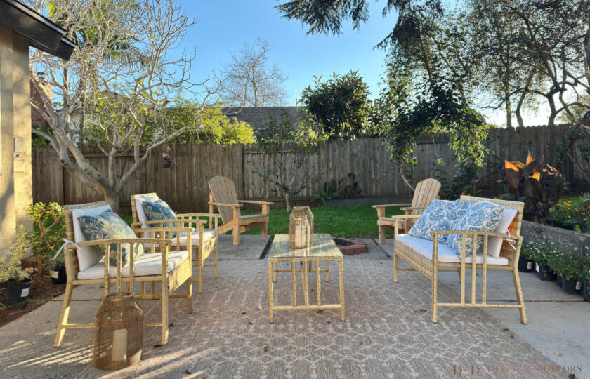 Staged outdoor patio living area featuring wicker patio seating, blue floral outdoor pillows, woven outdoor rug, metal lanterns, and a brick fire pit in the yard.