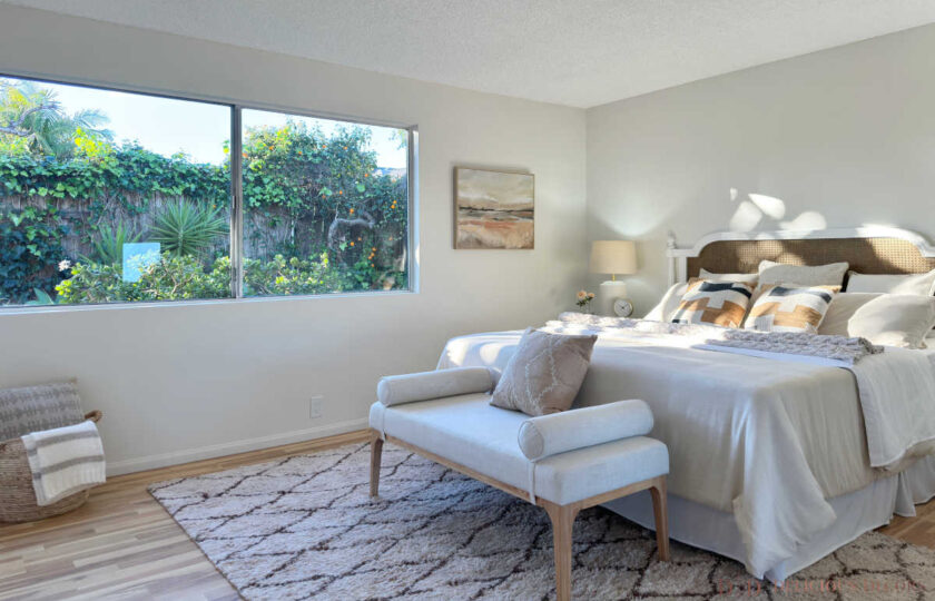 Spacious primary suite showing a large picture window facing lush green ivy foliage, diamond shag rug, bench, and cane headboard bed.