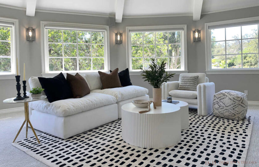 Curved sunroom lounge staged with white sectional, fluted drum coffee table, black-and-white grid rug, and bay windows.