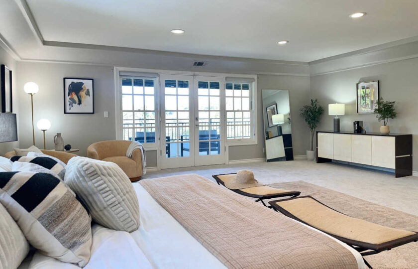 Luxury primary bedroom staged with white upholstered bed, dark nightstands, woven folding benches, and sitting area with armchairs.