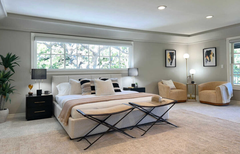 Luxury primary bedroom staged with white upholstered bed, dark nightstands, woven folding benches, and sitting area with armchairs.