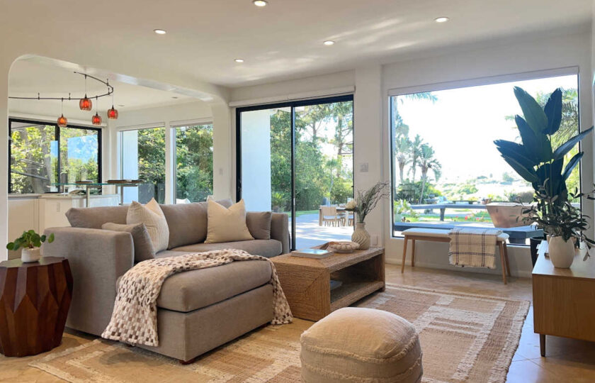 Open-concept Malibu living room featuring woven coffee table, grey sectional, textured pouf, and indoor plant styling.