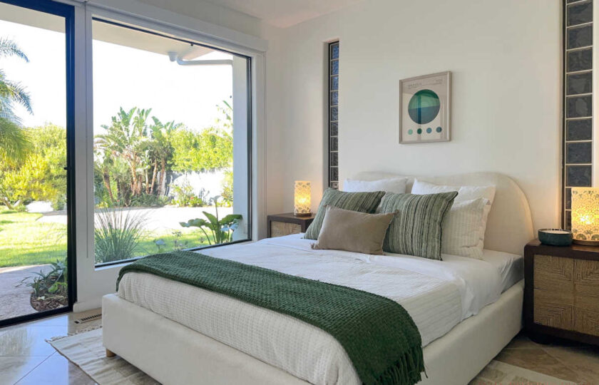 Staged Malibu bedroom with white upholstered bed, green throw blanket, woven nightstands, and floor-to-ceiling glass doors.