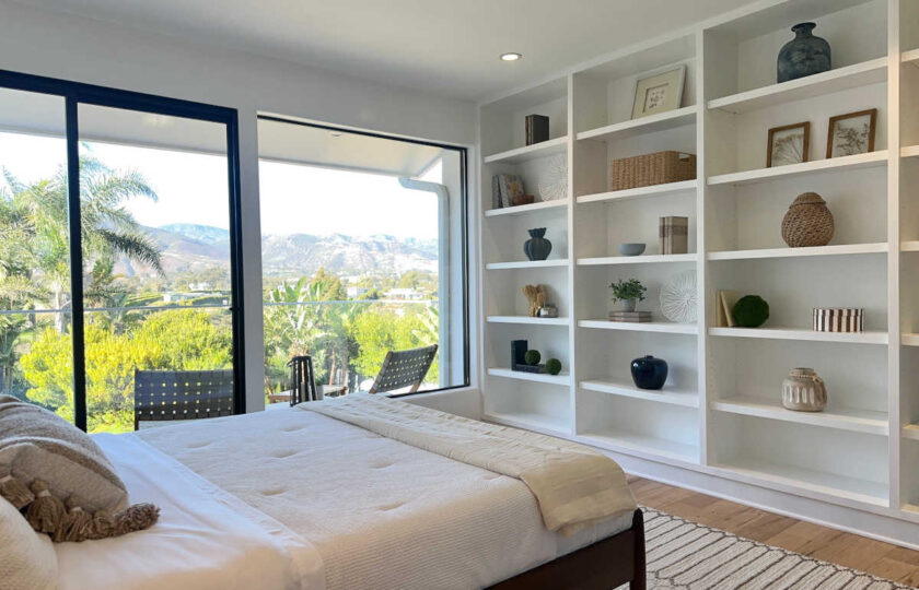 Staged bedroom in Malibu featuring mid-century wooden bed frame, custom white built-in bookcases, and balcony views.