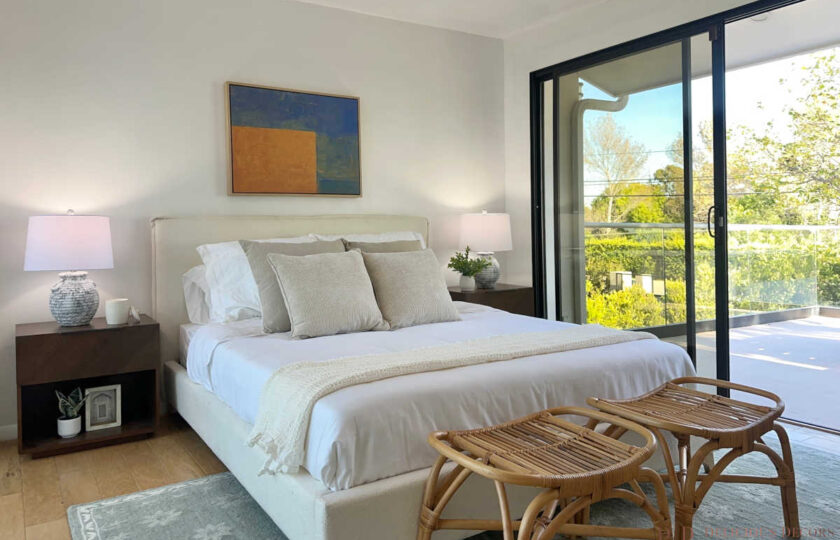 Malibu guest bedroom staged with cream upholstered bed, woven rattan stools, mid-century nightstands, and private balcony access.