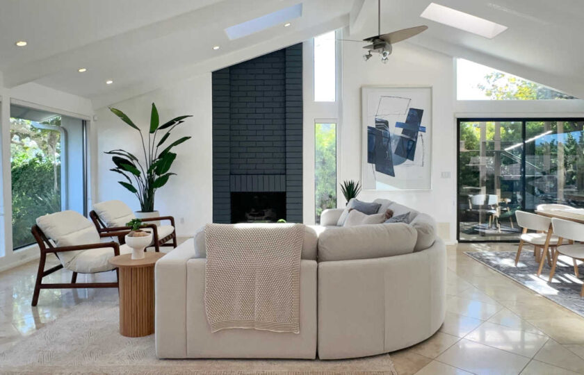 Vaulted great room staging in Malibu featuring dark brick fireplace, curved white sectional sofa, mid-century wood armchairs, and skylights.