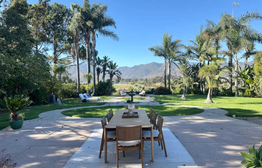 Staged Malibu estate patio with teak dining table, woven chairs, manicured lawn, and sweeping mountain backdrop.