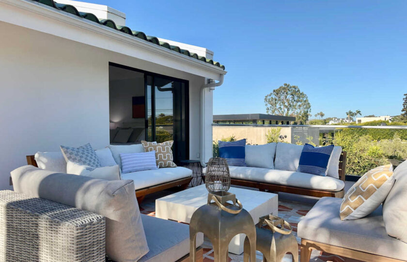Luxury ocean-view balcony staging in Malibu featuring teak outdoor sofas, blue accent pillows, and modern brass lanterns.